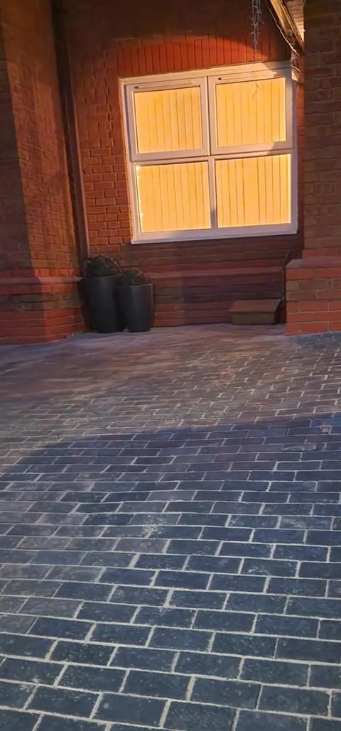 Brick building exterior with large window illuminated from within, black planters below, and patterned brick pavement