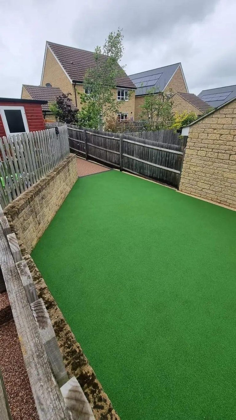 Residential backyard with bright green artificial turf surrounded by stone walls and wooden fences, backed by suburban homes with solar panels