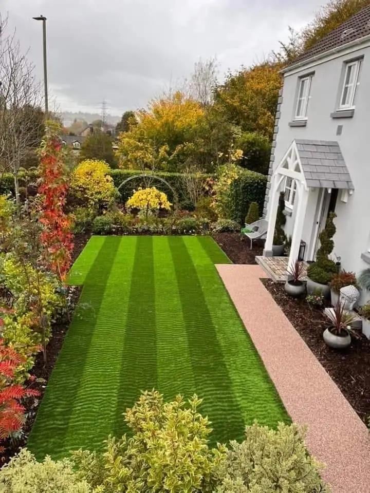 A well-maintained residential garden with striped green lawn, autumn foliage, and a white house with paved walkway
