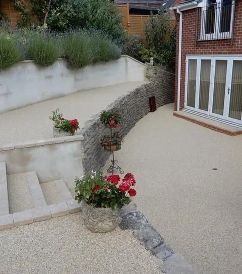 Modern residential patio with gravel surfacing, stone retaining walls, flowering planters, and brick house with white garage doors