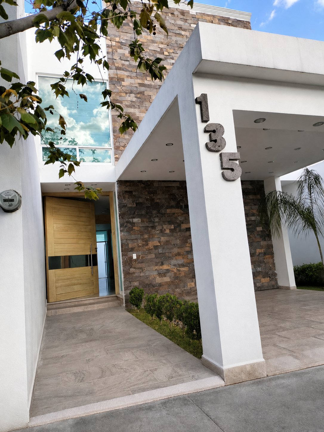 Fachada moderna de la casa con muros blancos, acabado de piedra decorativa, puerta principal de madera con vidrio, cochera techada y número 135