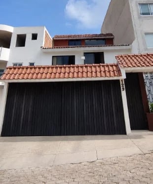 Fachada de la casa con muros blancos, techo de teja terracota, portón de cochera negro con lamas verticales, segundo piso con ventana de cristal, cielo azul con nubes blancas y piso de concreto