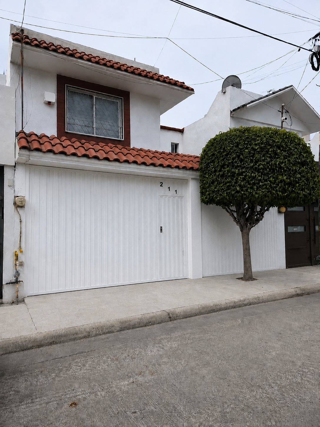 Fachada de casa de dos pisos color blanco con techo de teja roja, puerta de cochera blanca con número 211 y árbol en el frente