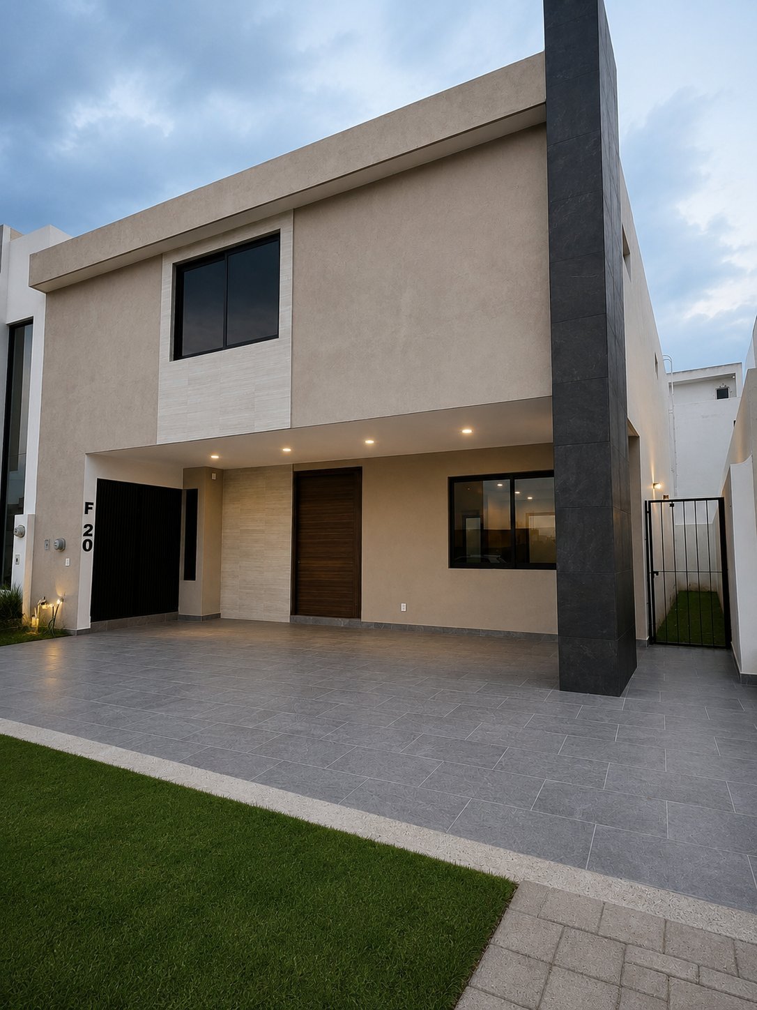 Fachada de la casa de dos pisos color beige con columna de acento oscuro, cochera de adoquín y número F 20 al atardecer