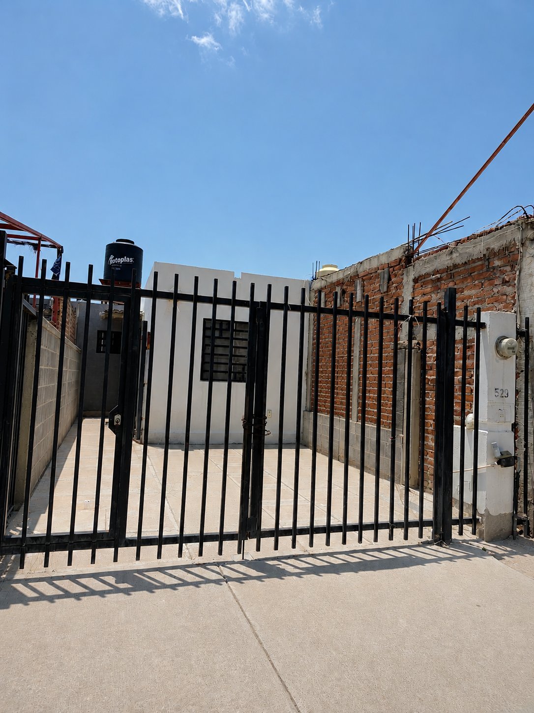 Fachada de la casa con portón de reja metálica negra, interfón, fachada de ladrillo y construcción blanca al fondo