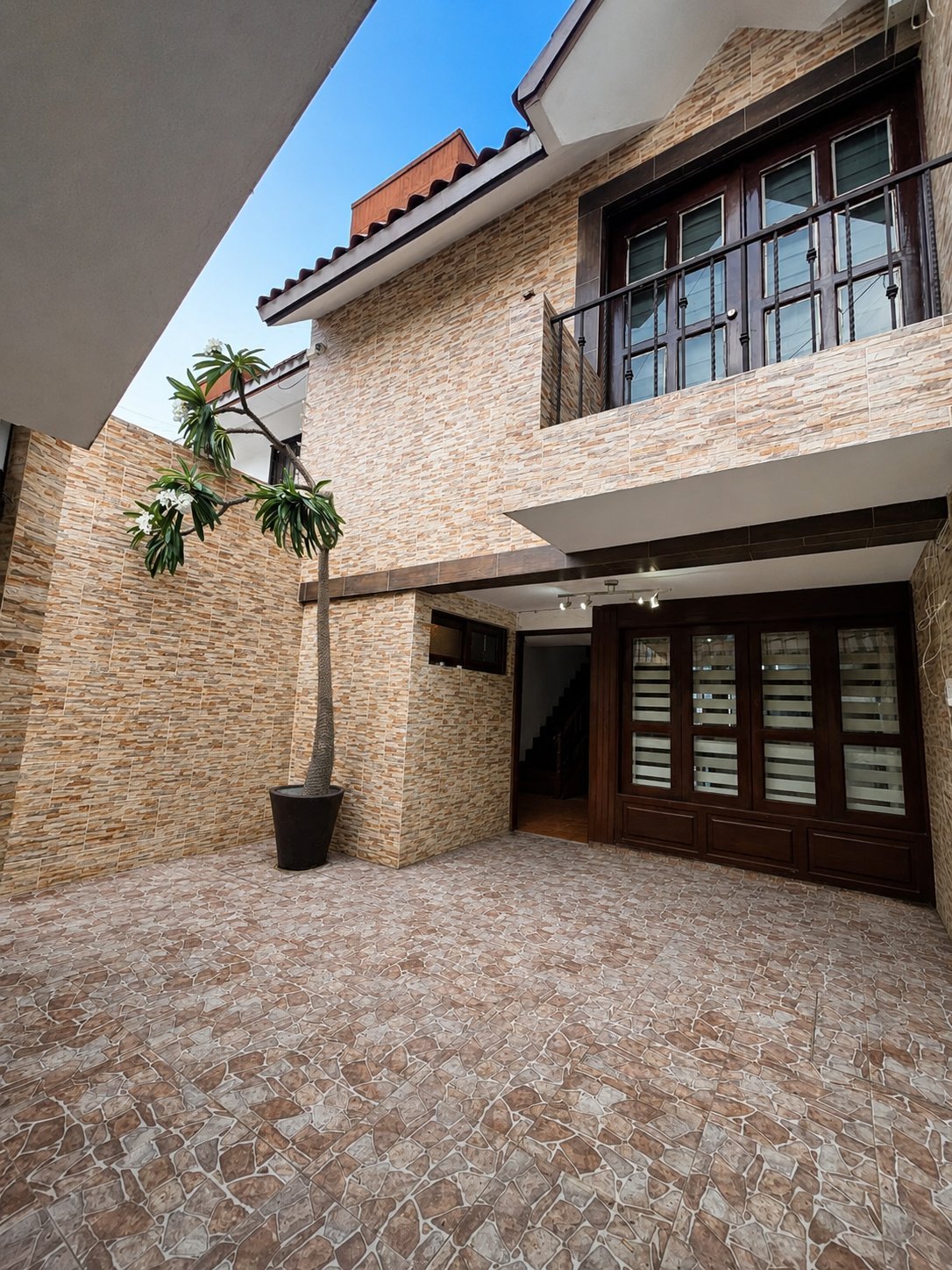 Fachada interior de la casa con patio de adoquín, planta de maceta, puerta de entrada de madera oscura con vitrales y balcón en planta alta