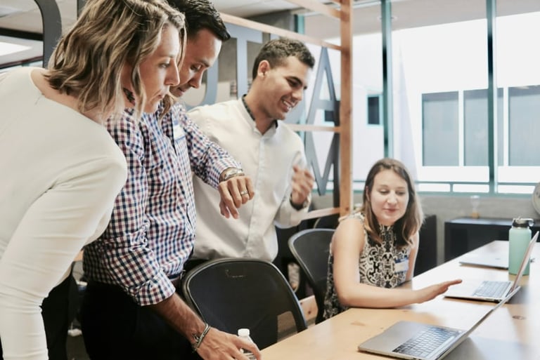 Four professionals gathered around a desk in a modern office, collaborating on a project with laptops and papers