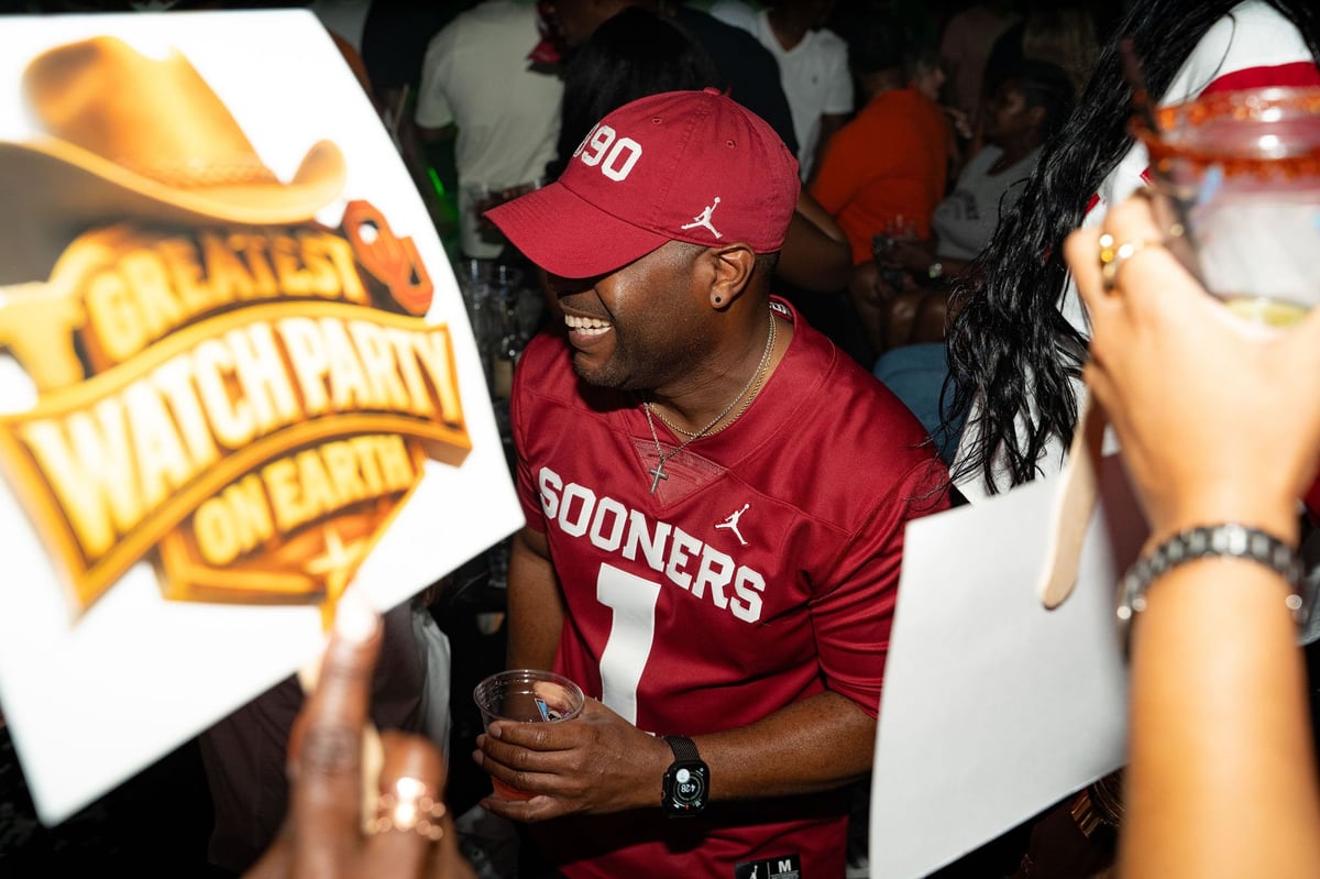 Man wearing red Sooners football jersey and cap at crowded event holding sign
