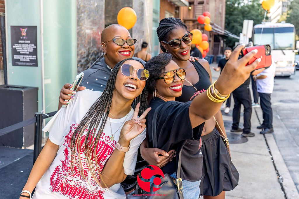 Four people taking a selfie on a city street, wearing colorful sunglasses and party attire with yellow balloons in background