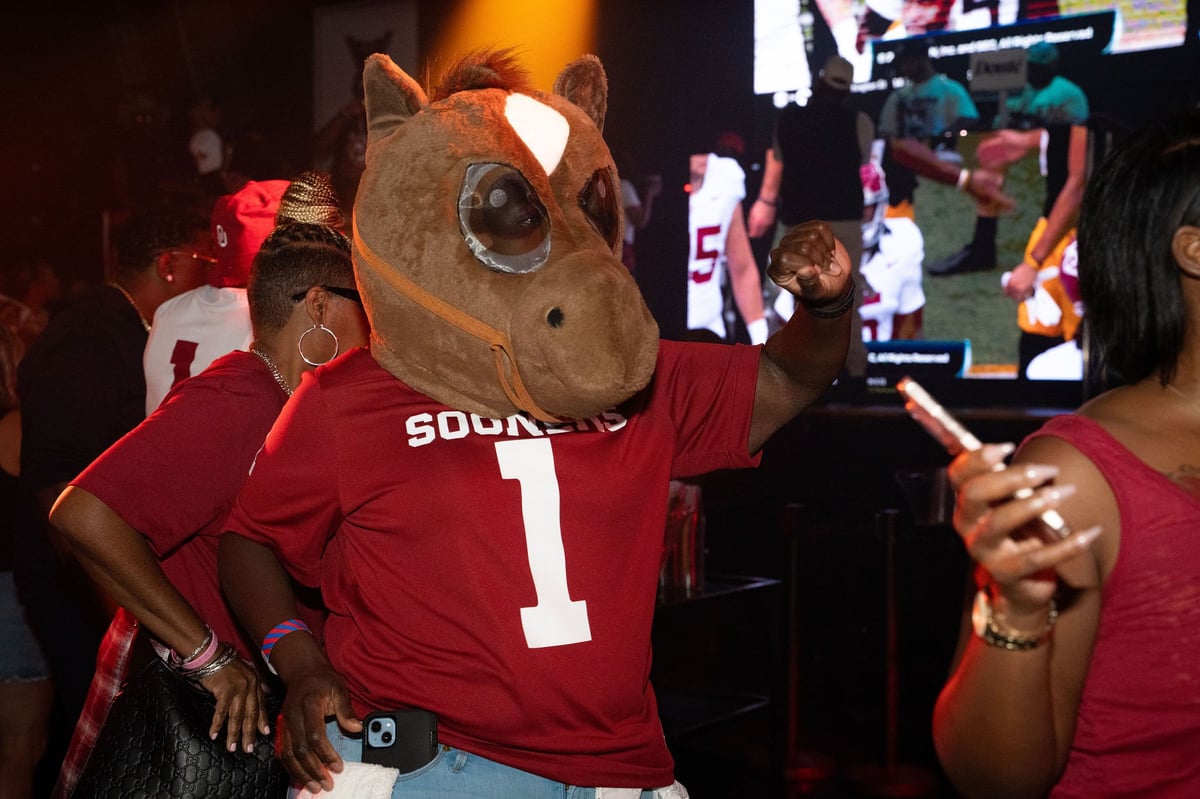 A mascot wearing a horse head costume and red number 1 jersey at a lively crowd event with a TV screen in the background