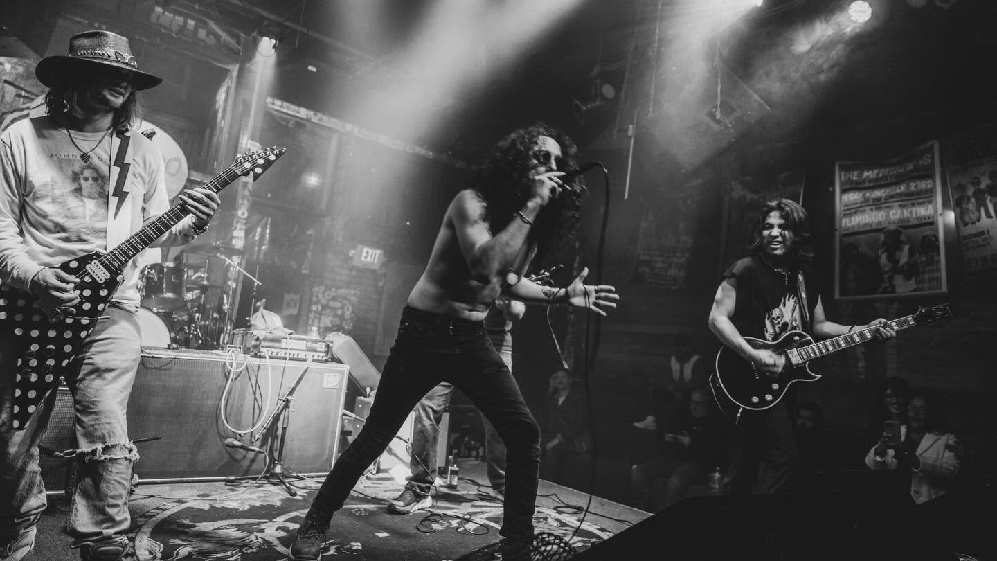 Black and white image of a rock band performing on stage with dramatic lighting, featuring guitarists and a lead vocalist