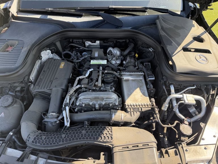 Open car engine bay showing a dark engine block with various mechanical components and hoses, viewed from above in daylight