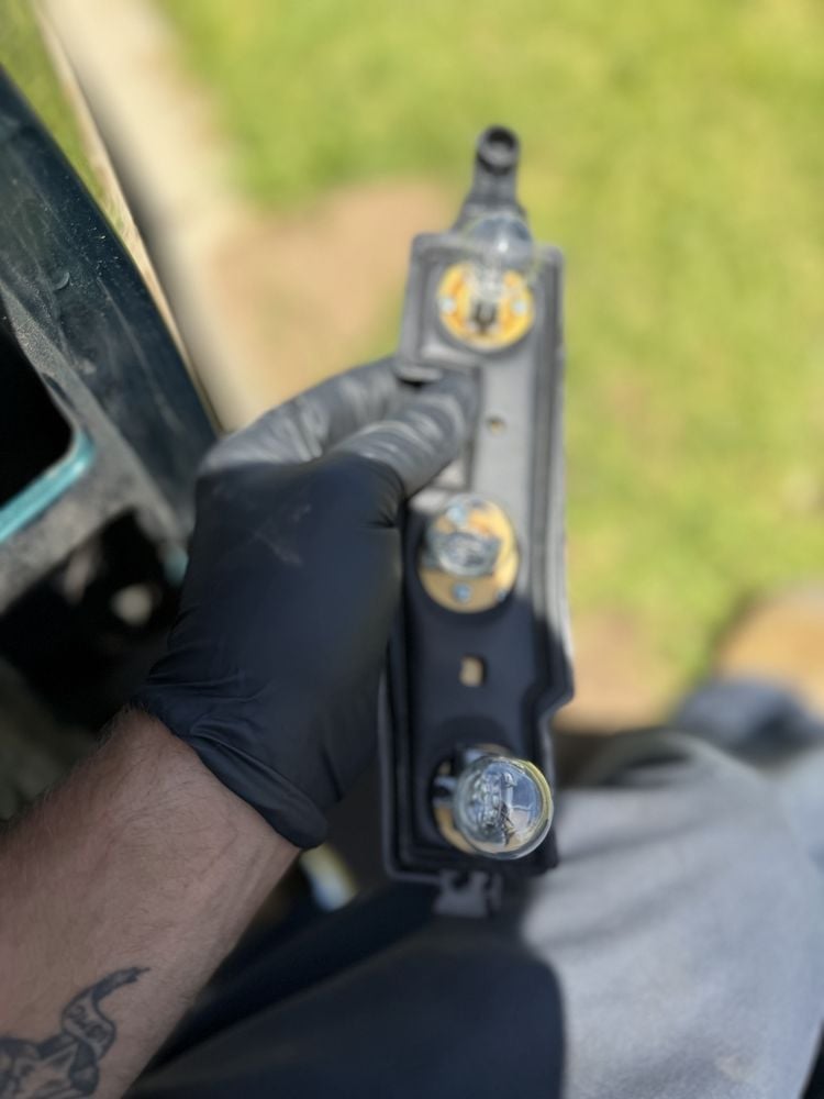 Close-up of a person's hand holding a black handgun with brass sights and trigger, outdoors with blurred green background