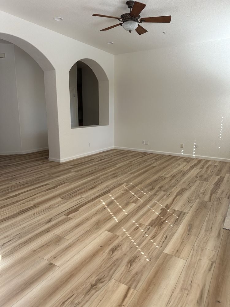 Empty room with light wood flooring, white walls with arched doorway, ceiling fan, and natural sunlight streaming through windows