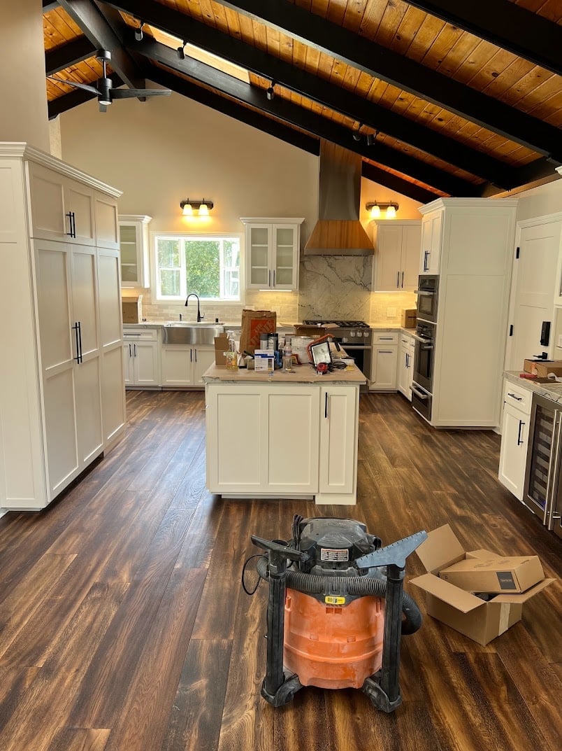 Modern kitchen with white cabinetry, wood beam ceiling, and work equipment on dark wood flooring during renovation
