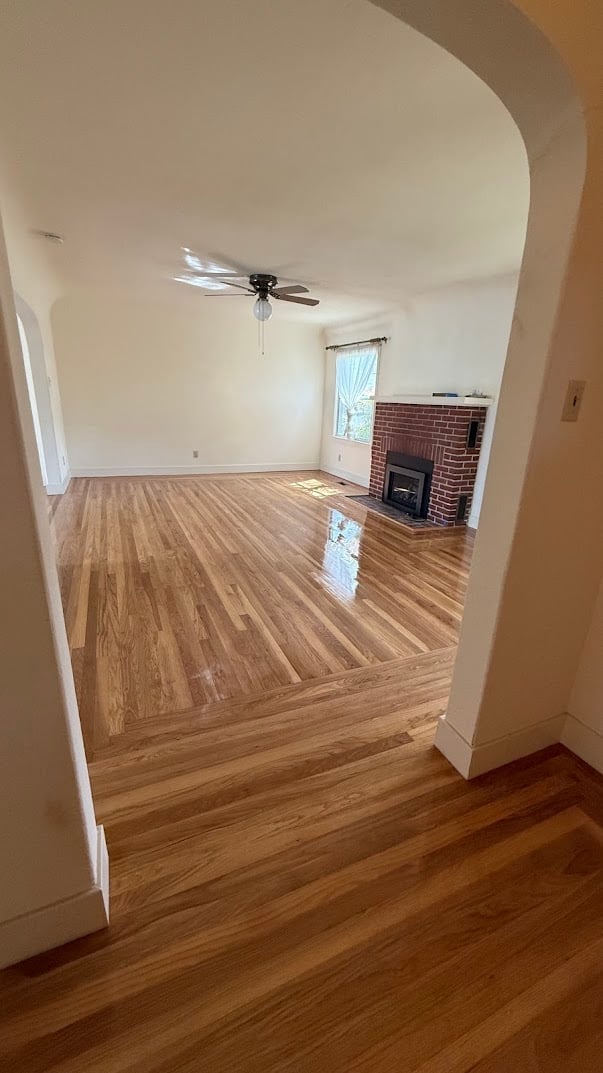 Empty living room with hardwood floors, ceiling fan, brick fireplace, and arched doorway entrance