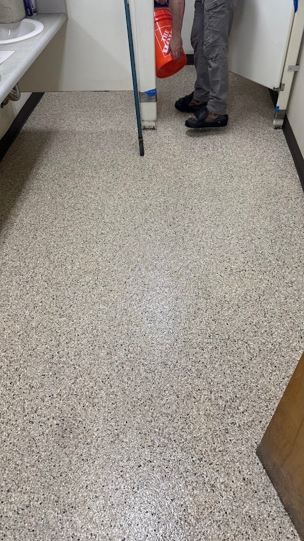 Epoxy coating being applied to a bathroom floor with speckled gray and white finish, person standing nearby with red bucket and blue painters tape on walls