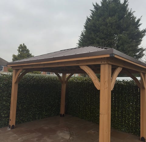 Wooden pergola structure with metal roof in backyard, flanked by evergreen hedges and concrete foundation