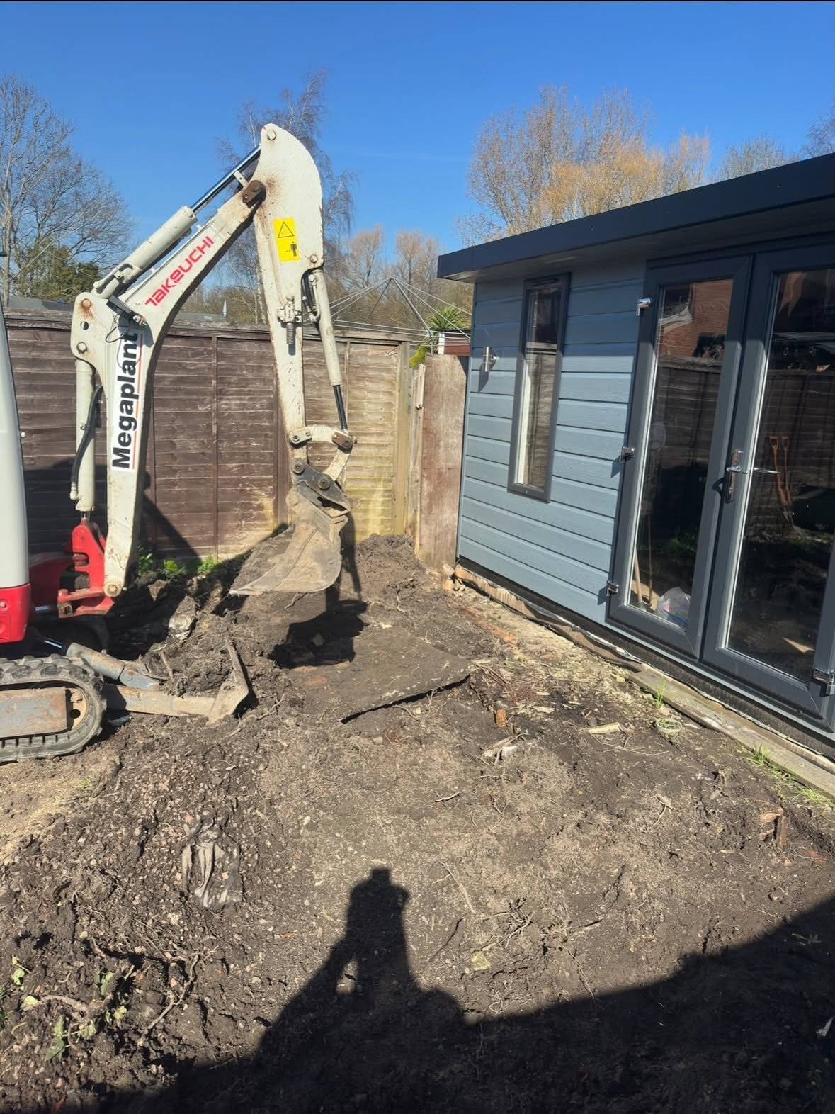 Mini excavator operating in backyard next to blue garden shed on dirt ground