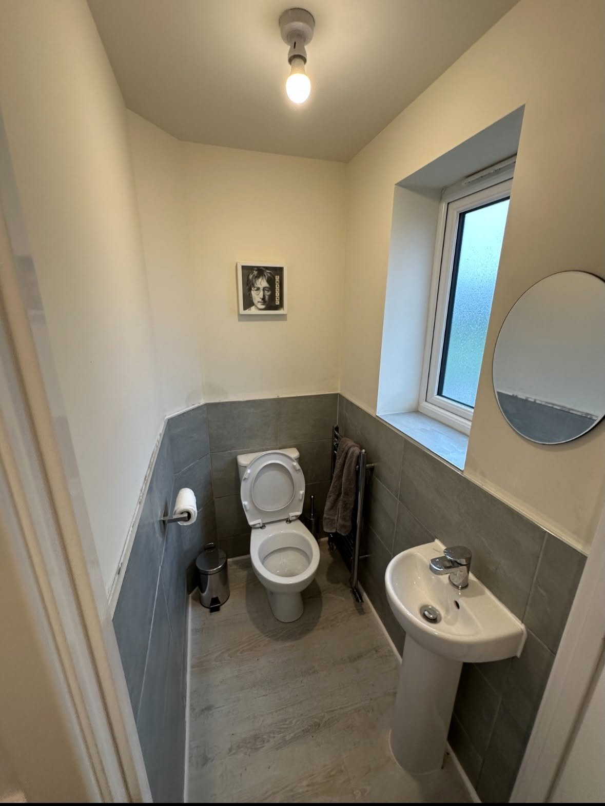 Small bathroom with cream upper walls, dark gray lower tile, toilet, corner sink, round mirror, and window with daylight