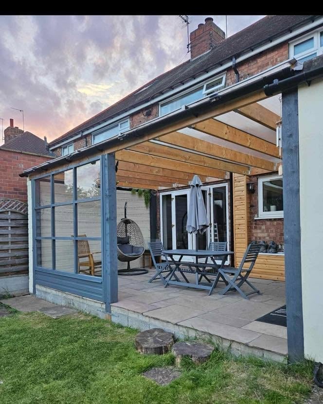 Modern blue pergola patio attached to brick house with wooden decking, outdoor furniture, and hanging chair at dusk