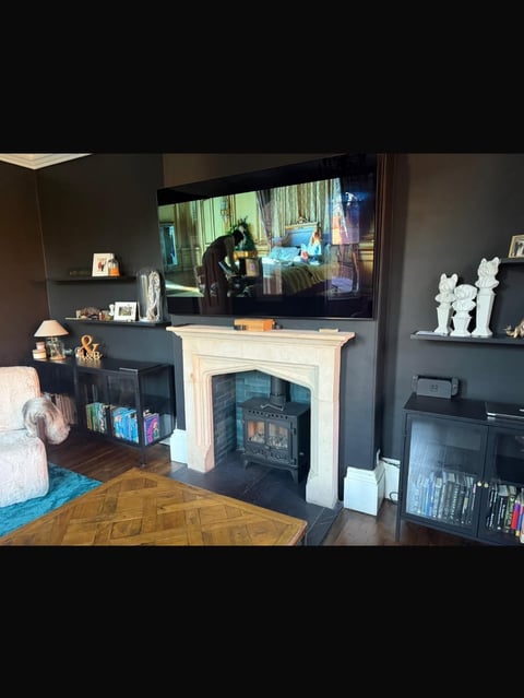 Modern living room with dark walls, flat-screen TV mounted above white fireplace with black stove insert, bookshelves, and decorative objects