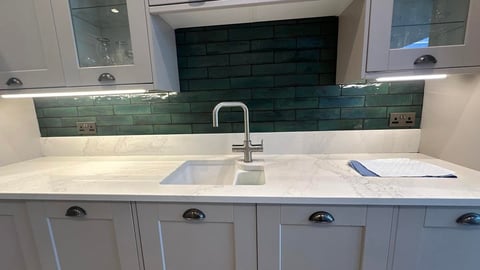 Modern kitchen sink with white countertop, chrome faucet, and dark green subway tile backsplash with upper cabinetry