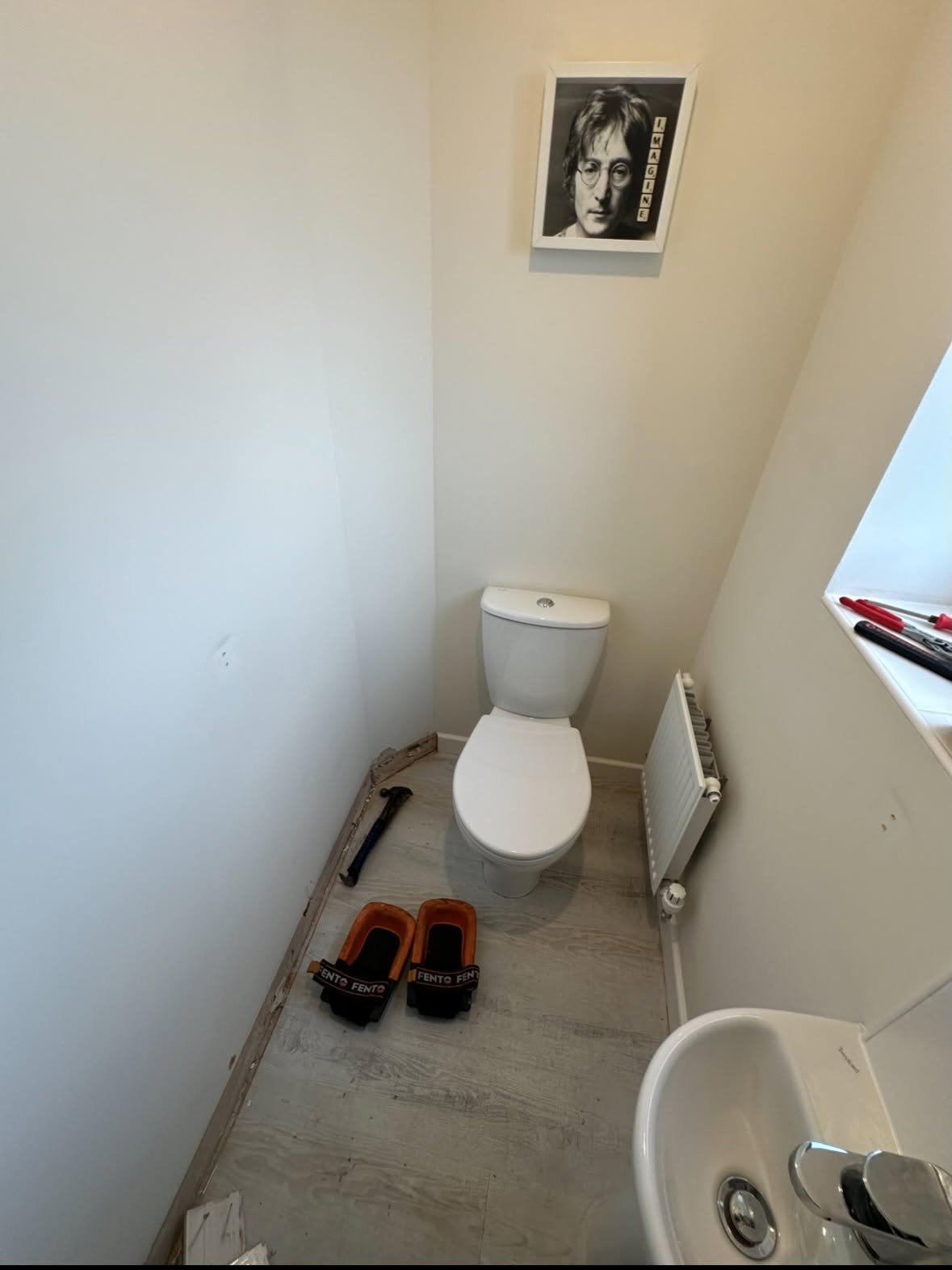 Small bathroom toilet with white fixtures, beige walls, a portrait poster above, and red slippers on the floor