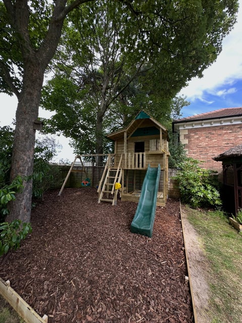 Wooden childrens playhouse with slide in a backyard shaded by large trees, with brick building visible in background