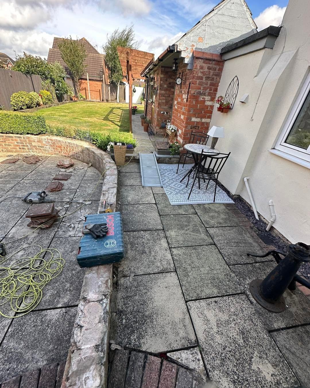 Residential garden patio under renovation with tools, equipment, and coiled garden hose on tiled floor beside brick cottage