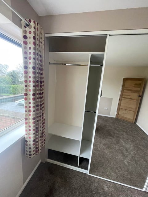 Empty bedroom with open mirrored wardrobe, patterned curtains, wooden door, and gray carpet