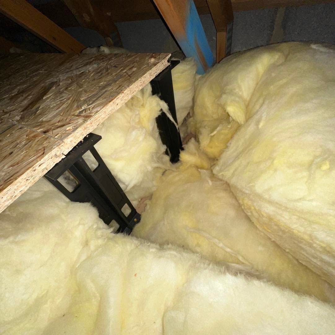 Yellow fiberglass insulation batts with black fasteners secured between wooden joists and a concrete beam in an attic space