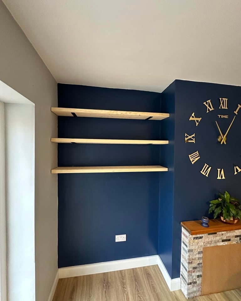 Modern home nook with navy blue accent wall, three wooden floating shelves, large gold Roman numeral clock, potted plant on wooden shelf, and hardwood flooring.