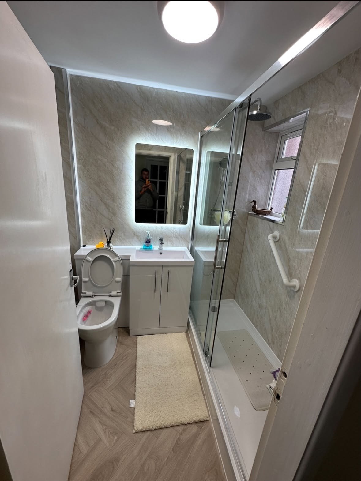 Modern bathroom interior with toilet, vanity sink, illuminated mirror, and glass shower enclosure with natural wood flooring