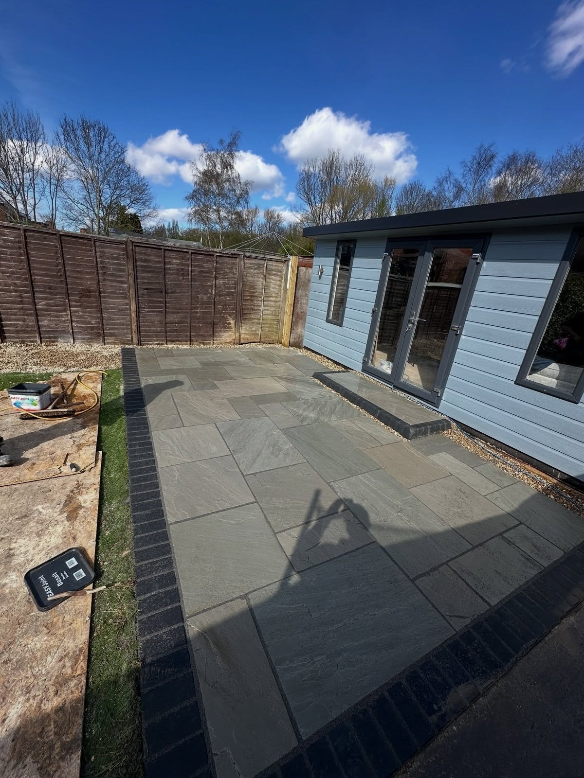 Newly paved patio with dark tiles next to a blue and white residential building with wooden fence, under clear blue sky
