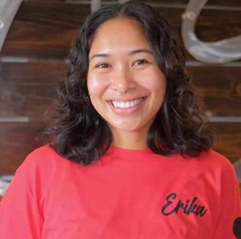 Smiling young woman with long dark wavy hair wearing a red shirt with text, standing indoors against a wooden background