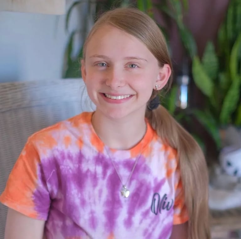 Smiling girl wearing a tie-dye purple and orange t-shirt, photographed indoors with potted plants in the background