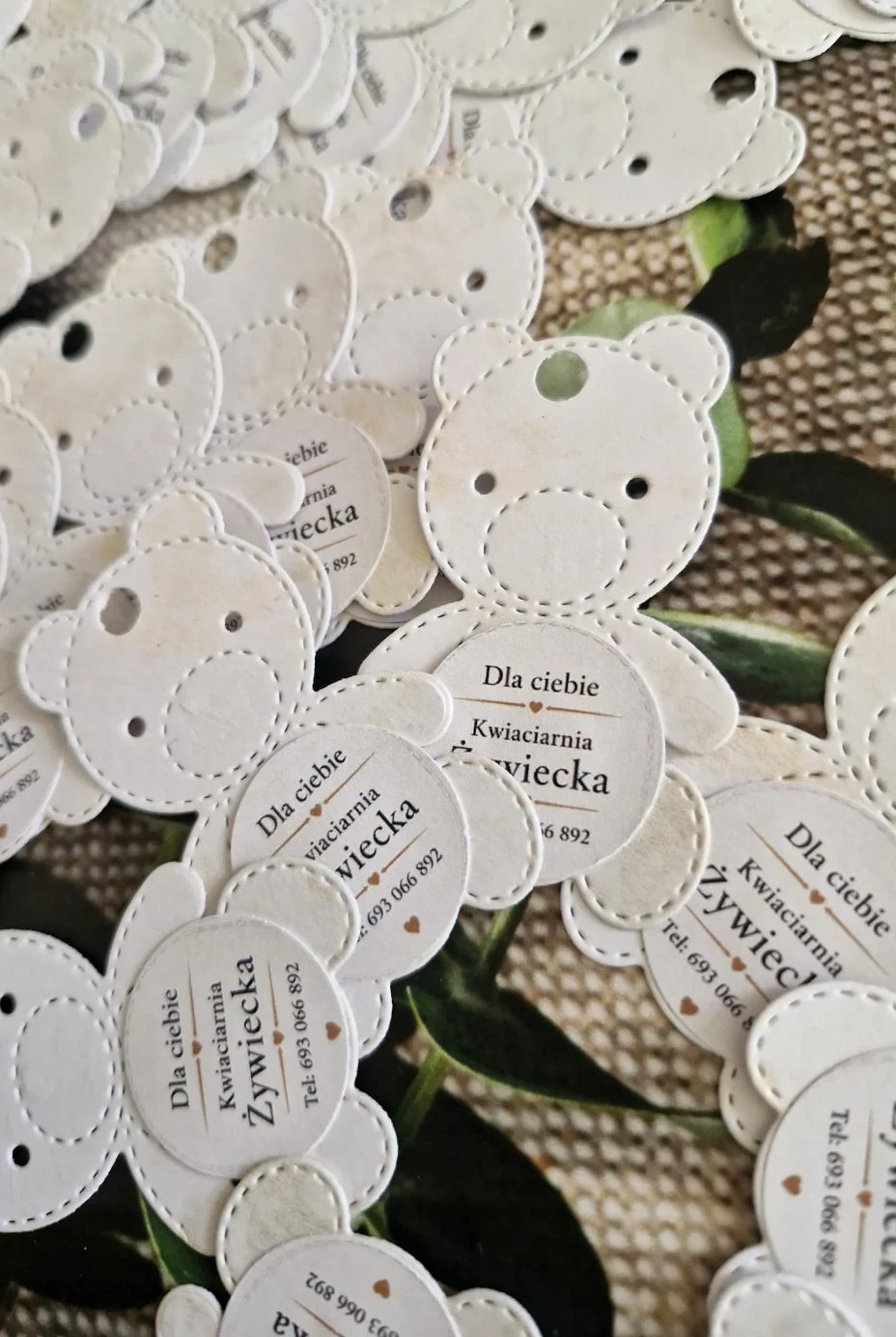 White paper gift tags shaped like teddy bears with Polish text printed on them scattered on a burlap surface