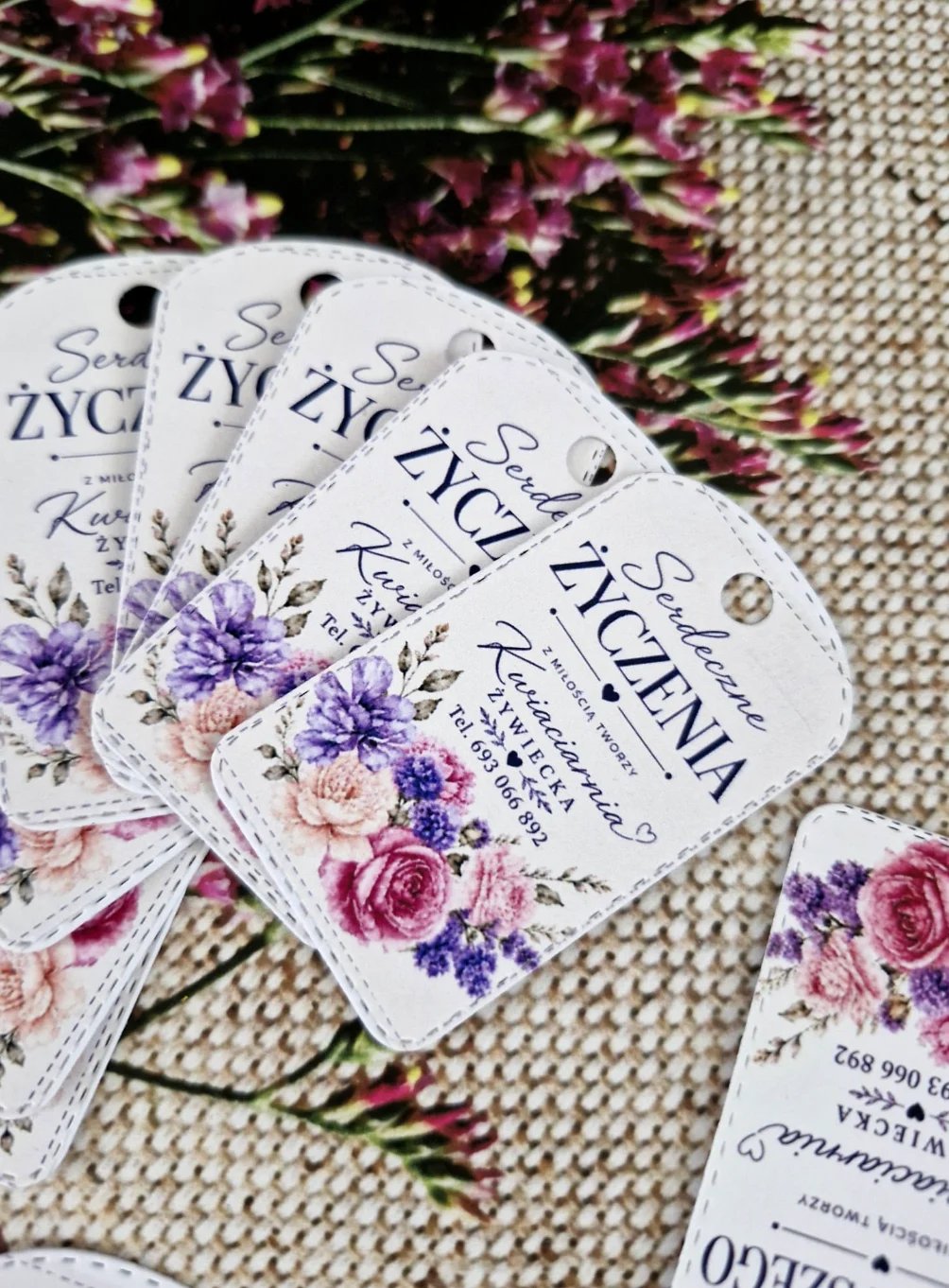Multiple product hang tags with floral designs featuring purple and pink flowers, arranged on a textured beige fabric with fresh pink and purple flowers
