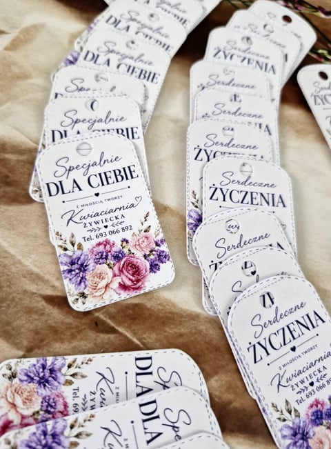 White gift tags with Polish text and purple floral designs arranged on beige fabric