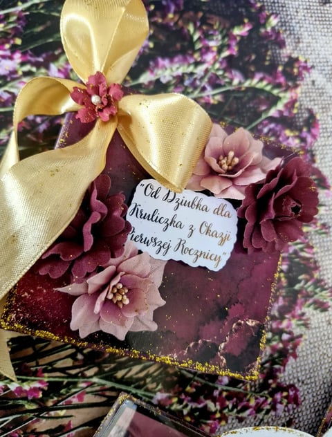 Decorative cake with burgundy and pink sugar flowers, gold ribbon bow, and handwritten message card on patterned surface