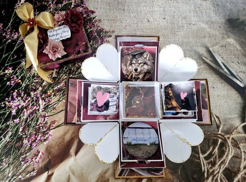 Handmade scrapbook layout with dried flowers, framed photos, white paper petals, and a gold ribbon on linen fabric