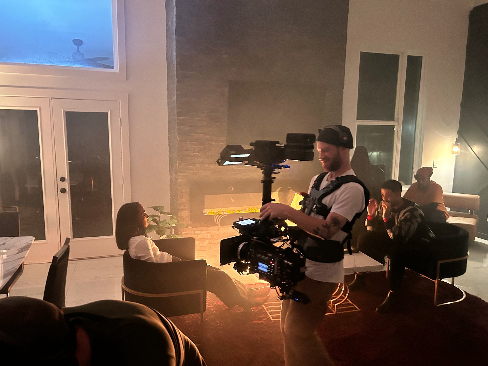 Cameraman filming an interview with a woman seated in a studio with a brick fireplace, while crew watches from a couch