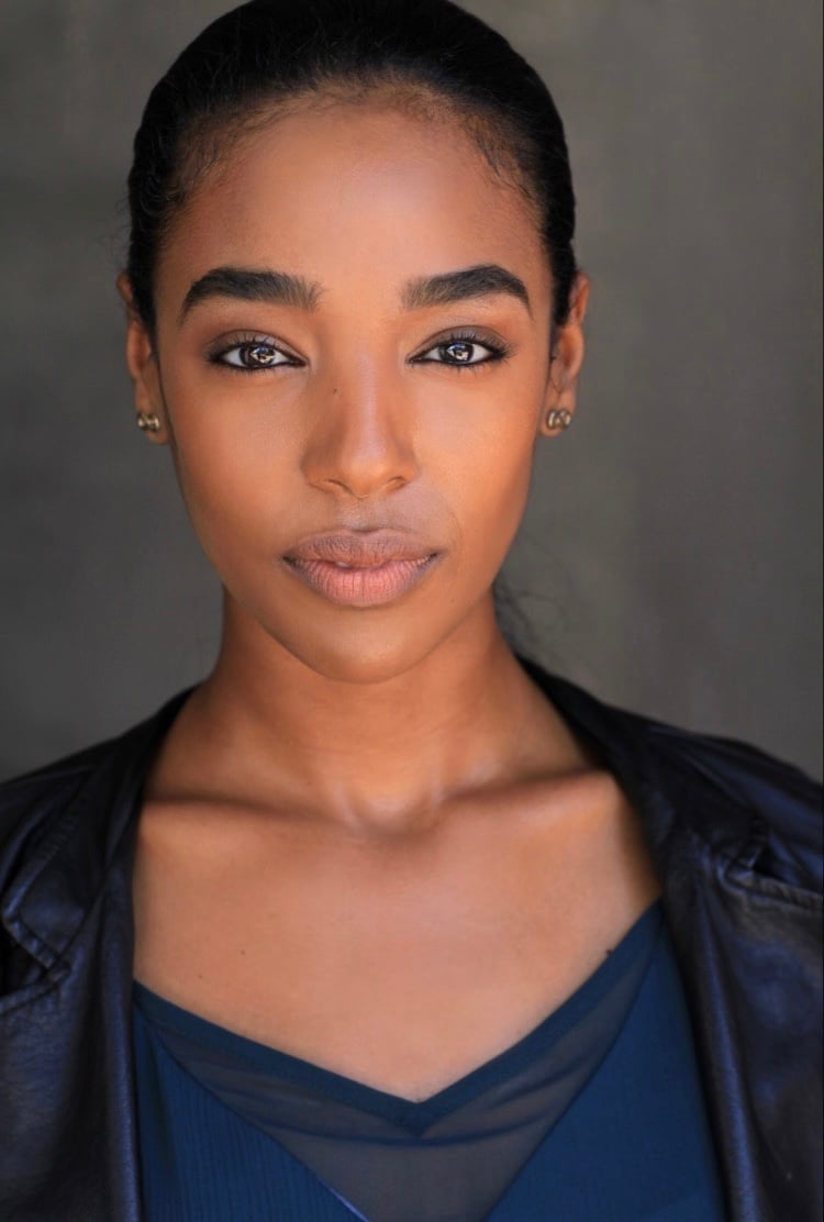 Professional headshot of Segen Gebremariam with dark hair pulled back, wearing a dark blue v-neck top against a gray background