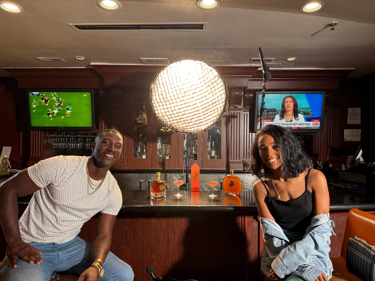 Two people sitting at a bar counter with drinks, large white spherical lamp hanging above, TV screens on walls