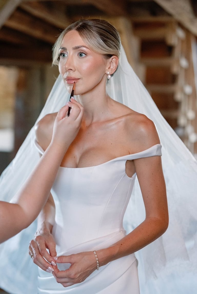 Bride in white off-shoulder wedding gown and veil applying lipstick while looking to the side