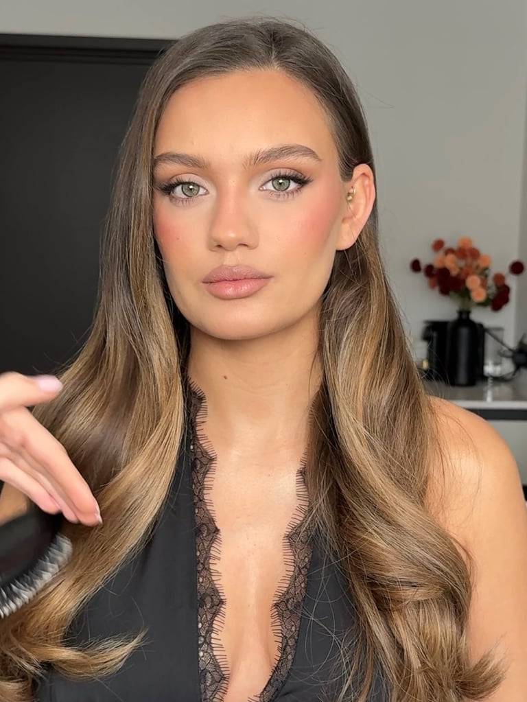 Woman with long caramel-toned hair and warm makeup looking at camera in indoor setting