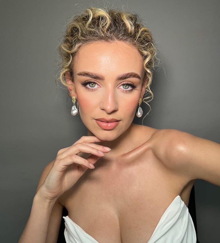Portrait of a woman with curly blonde hair, pearl earrings, and pink lipstick against a gray background