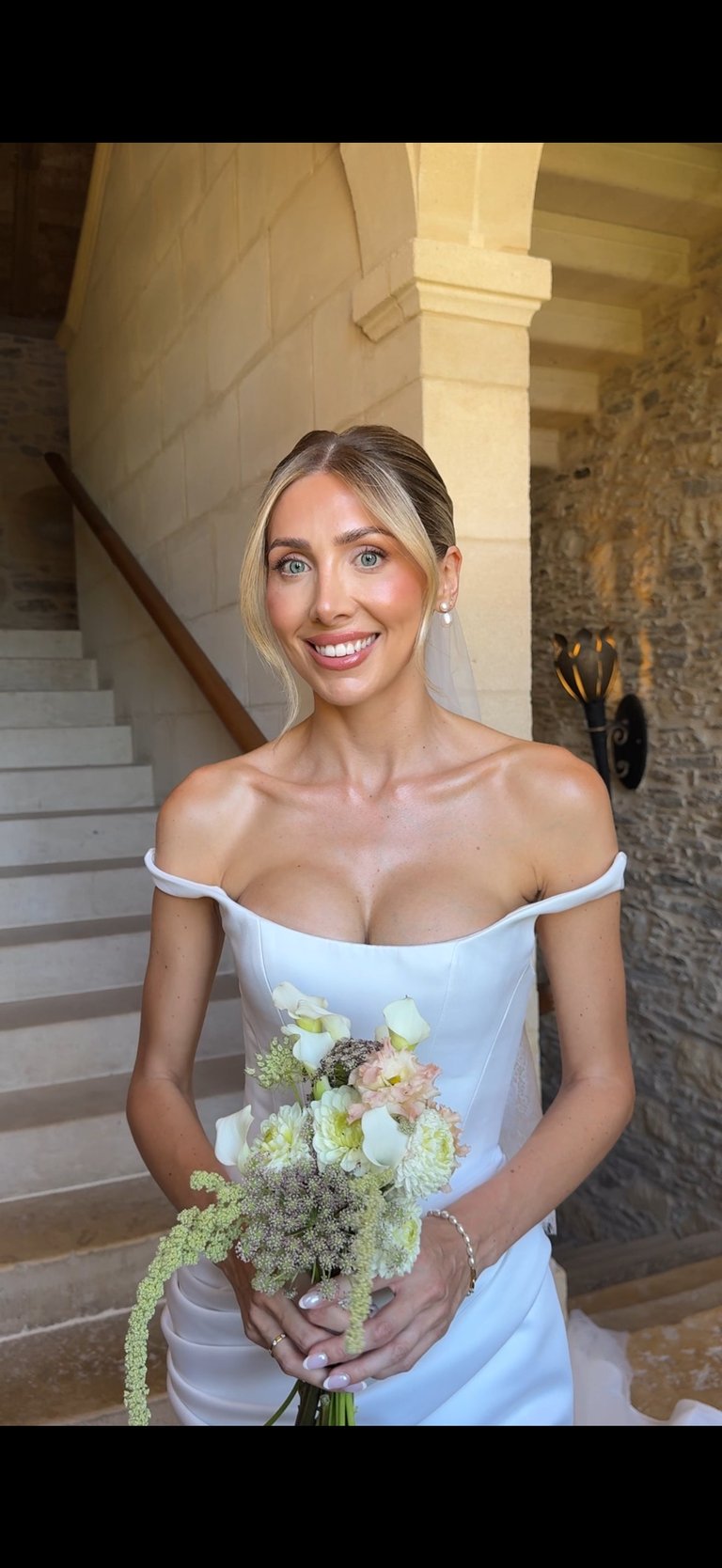 Woman in white off-shoulder dress holding pastel wedding bouquet, smiling at camera in elegant stone corridor