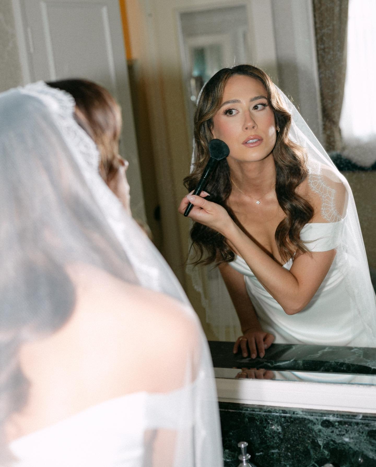 Woman with long dark hair applying makeup while looking in mirror, wearing white off-shoulder top and sheer veil